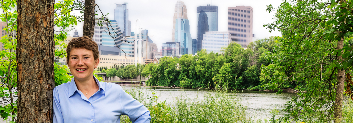 Astrid Bryce Header Image - Astrid leaning  against a tree on the green banks of the Mississippi River in front of the Minneapolis skyline
